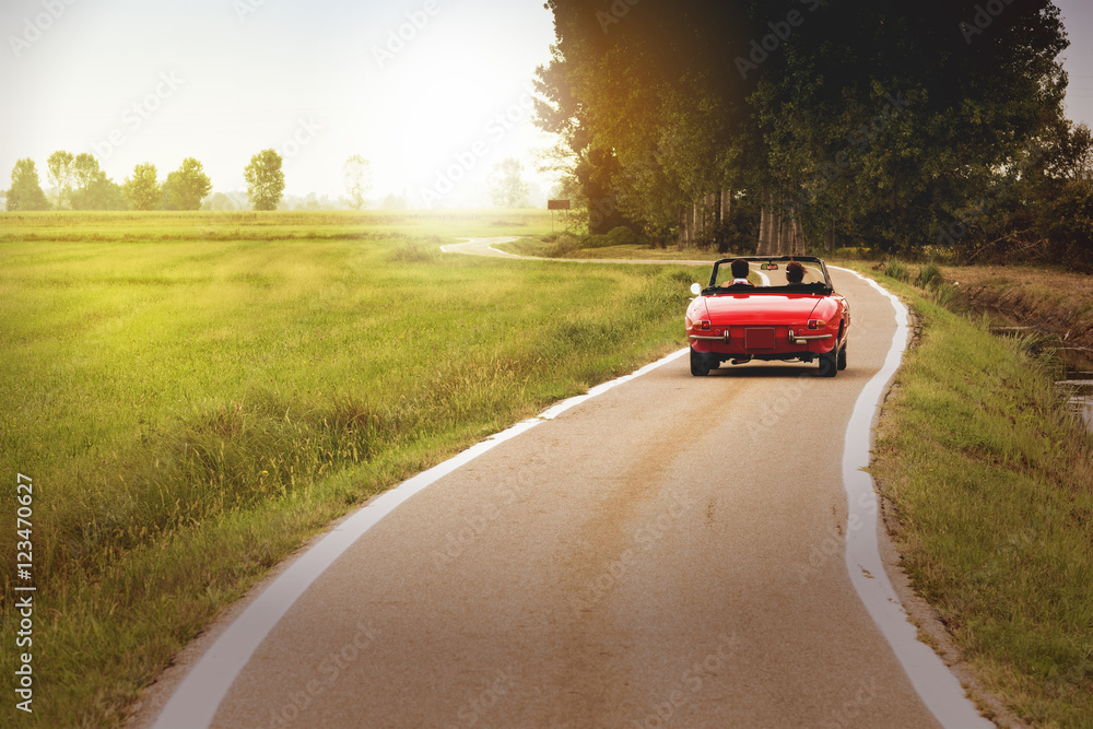 Classic red convertible car traveling in the countryside at sunset - obrazy, fototapety, plakaty 