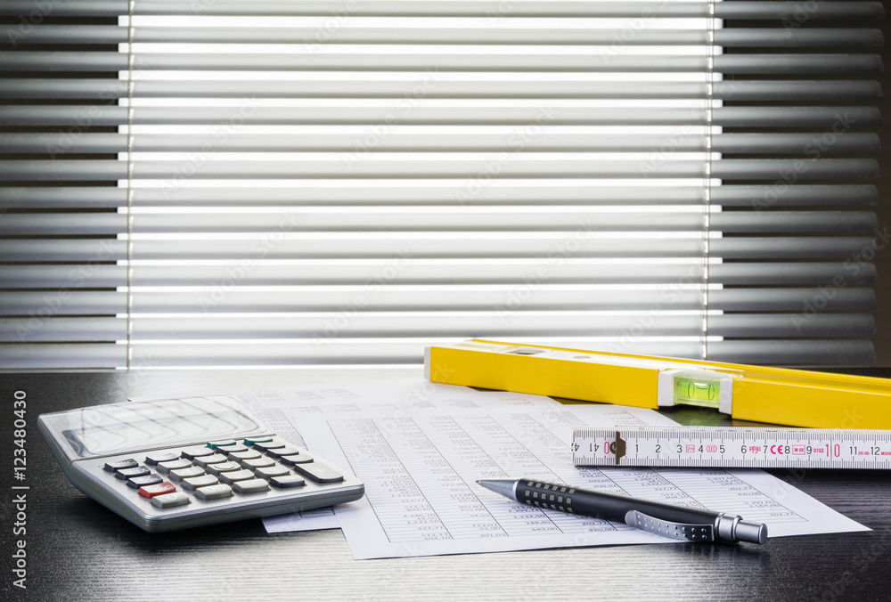Kugelschreiber, Tabellen, Taschenrechner, Zollstock und Wasserwaage auf Schreibtisch vor Fenster mit Jalousie