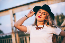 Outdoor Lifestyle Close Up Portrait Of Happy Blonde Young Woman In Stylish Casual Red Dress (skirt) And Black Hat  Stay On Bridge On The Street. Pretty Hipster Girl Having Fun And Enjoying Holidays.
