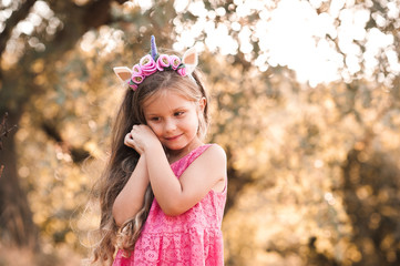 Smiling baby girl 4-5 year old wearing unicorn hairband and pink dress outdoors. Childhood.