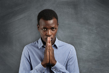 Portrait of handsome dark-skinned student holding hands in prayer, looking worried and impatient, anticipating results of final exams or begging teacher to give him another chance. Body language