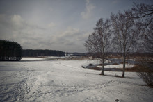 River Runs Behind A Field Covered With Snow Under Grey Winter Sk