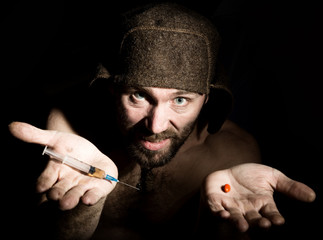 Wall Mural - Dark portrait of scary evil sinister bearded man with smirk, offers a variety of drugs, a syringe or a pill. strange Russian man with a naked torso and a woolen hat