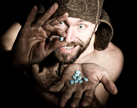 Dark portrait of scary evil sinister bearded man with smirk, holds in palm of a bunch of pills. strange Russian man with a naked torso and a woolen hat