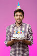 Young handsome man smiling, holding birthday cake over purple background.