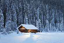 A Snow Covered Log Cabin On A Snow Covered Lakeshore Surrounded By Evergreen Trees At Dusk;Lake Louise Alberta Canada