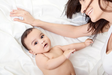 Wall Mural - Young mother playing with her baby on white silk bed sheet. Cute