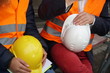 © cineberg - Cropped construction workers wearing safety vest holding hardhats
