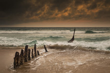 Wooden Posts On A Beach With A Boat Being Tossed In The Water And Waves Crashing Into The Sand Under A Cloudy Sky; Queensland, Australia