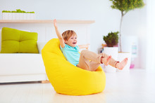 Excited Kid Having Fun, Sitting On Yellow Bean Bag At Home