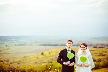 Wall Mural - Wedding couple poses with green paper hearts on the hill with gr