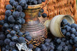 Old wine pitcher and clay glass surrounded by black grapes in a wicker basket with metal winemaking emblem and a cork.