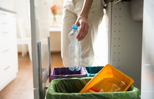 Woman Putting Empty Plastic Bottle In Recycling Bin In The Kitchen. Person In The House Kitchen Separating Waste. Different Trash Can With Colorful Garbage Bags.