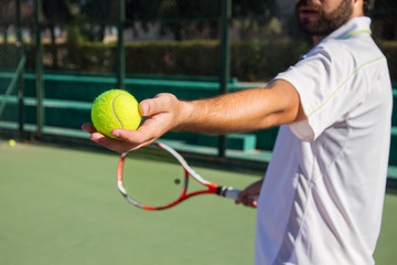 Wall Mural - Professional tennis player, bearded young man offering tennis ball to play the game. He has a racket in her right hand and wears a white cap. 