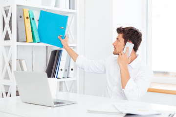 Poster - Young businessman taking folder from shelf and talking on phone