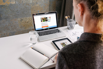 Wall Mural - Back view. Business woman in gray shirt sits at white desk.On table is 
laptop,on-screen page online store,near notebook,digital tablet,glass of
 water and pencil holders.Woman working in office.