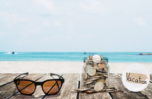 Saving Money For Vacation Concept, Glass Coins Container With Sunglasses On Wooden Table At Tropical Beach
