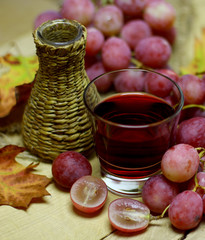 Red rose grapes bunch, wicker bottle, glass with red natural organic house wine or juice, and autumn leaves on wood background.