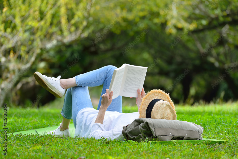 Happy woman lying on green grass reading a book in the park (outdoors) - obrazy, fototapety, plakaty 