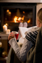 Woman Is Sitting With Cup Of Hot Drink And Book Near The Fireplace