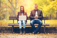 Young Couple Sitting Ona A Bench In A Park On A Beautiful Autumn Day. She Is Using Laptop And Young Man Is Using His Mobile Phone.