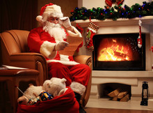 Happy Santa Claus Sitting At His Room At Home Near Christmas Tree And Big Sack And Reading Christmas Letter Or Wish List