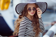 Beautiful Woman Model Looks,brunette With Long Curly Hair,wears Dark Sun Glasses On His Head Wearing Striped Straw Hat With A Large Brim,dressed In A Striped Summer Dress,spends Time On The Ocean