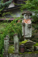 Takayama, Japan - September 24, 2016: Bodhisattva stone statue in garden of Hikakokubun-ji Buddhist Temple. Lots of green foliage and moss. Idol has red apron and a dome hat.