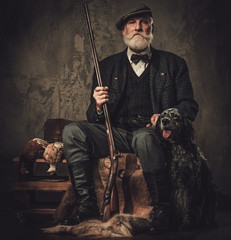 senior hunter with a english setter and shotgun in a traditional shooting clothing, sitting on a dar