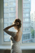 © natavilman - beautiful girl with long brown hair in a white dress standing at the window from the back and corrects hairstyle
