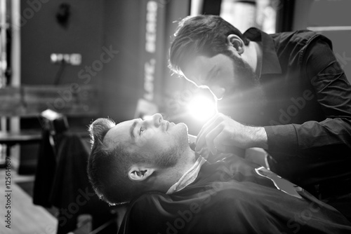 Fotografie, Obraz The Barber man in the process of cutting the beard of a client in a Barbershop,