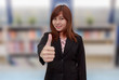 © petzshadow - Smiling businesswoman holding thumps up in font of book shelf