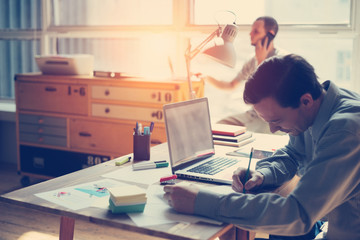 Fototapete - Man working on laptop in open space office. Paperwork on the table and manager talking on the phone