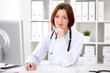 © rogerphoto - Young brunette female doctor sitting at a desk and working on the computer at the hospital office.  Health care, insurance and help concept. Physician ready to examine patient