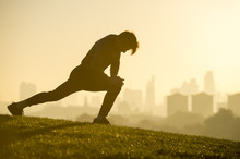 Male Jogger Stretching On The Grassy Top Of Primrose Hill In Front Of A Misty Golden Sunrise View Of The London City Skyline