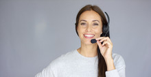 Adorable And Beautiful Female Call Center Agent Speaking With Someone On Headset. She Has Smile On Her Face. Isolated On Gray.