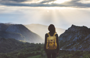 Hipster young girl with bright backpack enjoying sunset on peak of foggy mountain. Tourist traveler on background valley landscape view mockup. Hiker looking sunlight flare in trip in northern spain