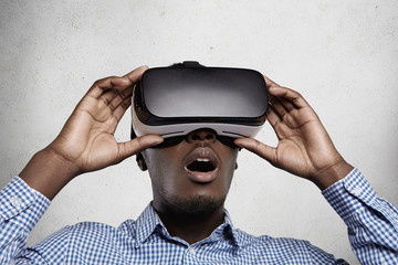 Wall Mural - Cyberspace, technology and entertainment concept. Amazed and fascinated dark-skinned office worker experiencing virtual reality using 3d headset glasses, opening his mouth widely with surprise