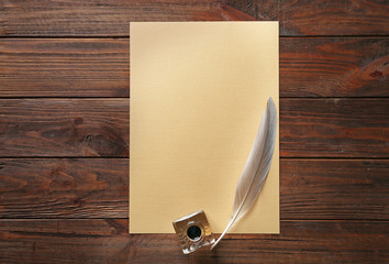 Inkwell with feather and sheet of paper on wooden background