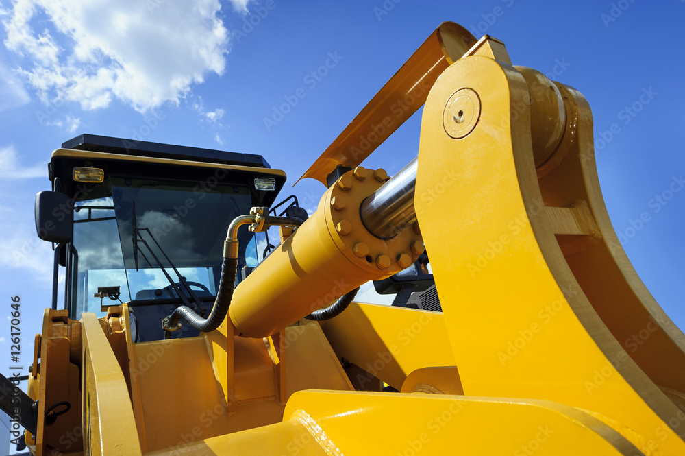 Bulldozer, huge yellow powerful construction machine with big scoop, focused on hydraulic piston arm, heavy industry, blue sky and white clouds on background 