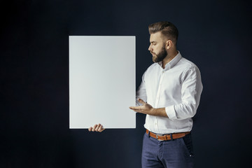 Young bearded businessman, dressed in a white shirt standing and holding a blank white poster. In the background dark blue wall. Mock up. Advertising poster. Ad.