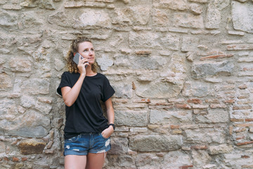 Wall Mural - Front view of a young woman dressed in denim shorts and a black t-shirt, standing and talking on the phone. In the background is bright stone wall. Girl uses a digital gadget.