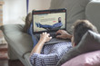 © GENETTICA - Young man lying comfortably on the sofa while using the laptop with series streaming website on the screen. View from behind. All screen graphics are made up.