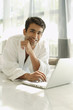 © Alexander - Man in bathrobe, using laptop, hand on chin, smiling at camera