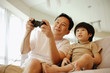 © Alexander - Father and son sitting on sofa, playing with video game, low angle view