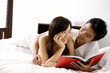 © Alexander - Couple lying on bed, holding book, smiling at each other