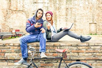 Teenage couple using phone and notebook outdoor at winter time - Best friends looking internet content sitting on old urban wall  - Concept of young people lifestyle with modern technologies and trend