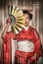 Asian Woman In Traditional Red Kimono Holding A Paper Fan, Which Covers Half Of Her Face.