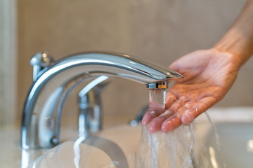 Woman taking a bath at home checking temperature touching running water with hand. Closeup on fingers under hot water out of a faucet of a sink or bathtub in house bathroom