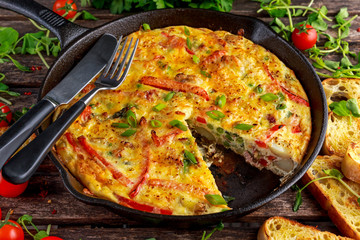 Frittata made of eggs, potato, bacon, paprika, parsley, green peas, onion, cheese in iron pan. on wooden table.